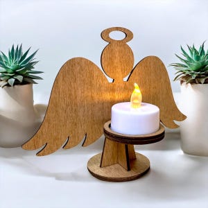 May include: Wooden angel-shaped candle holder with a battery-operated LED candle. The angel has outstretched wings and a halo. The candle is white with a flickering yellow light. Two potted succulents flank the angel.