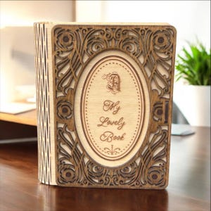 Puede incluir: Una caja de madera con forma de libro con un diseño intrincado cortado con láser. La caja está cerrada y tiene un acabado marrón claro. La parte delantera de la caja tiene un marco ovalado decorativo con el texto "My Lovely Book" grabado en el interior.
