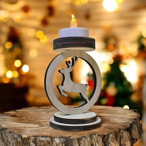 Puede incluir: Un portavelas navideño de madera con la silueta de un reno dentro de un marco circular. El portavelas tiene una vela blanca con una llama amarilla parpadeante. La base es de madera y está sobre una superficie de madera.