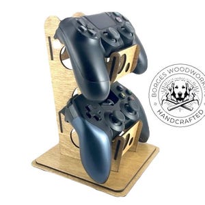 May include: Wooden gaming controller stand holding two black controllers. The stand is made of light brown wood and features a circular logo with the text "BORGES WOODWORKING HANDCRAFTED".