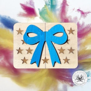 Può includere: Una scatola regalo in legno con un fiocco blu e stelle dorate. La scatola è in legno chiaro e ha una finitura naturale. Il fiocco è fatto di un unico pezzo di legno ed è tagliato a forma di fiocco. Le stelle sono anch'esse in legno e sono tagliate a forma di stella semplice.