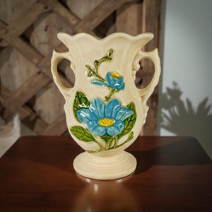 May include: Cream-colored ceramic vase with two handles and a pedestal base. The vase is decorated with blue flowers, green leaves, and yellow centers. The vase has a scalloped rim and is likely vintage.