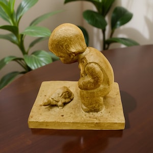 May include: A tan-colored sculpture of a child looking down at a turtle. The child is kneeling on a square base, with the turtle positioned in front. The sculpture has a matte finish and is set against a blurred background.