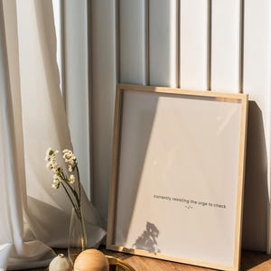 May include: A framed print with the text "currently resisting the urge to check" leans against a white wall. A gold tray holds a wooden sphere, a small decorative object, and a vase of white flowers. The scene is lit by natural light.