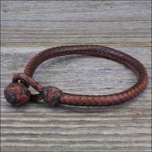 May include: Braided brown leather bracelet with a knotted closure and a decorative ball. The bracelet is made of interwoven leather strips, showcasing a rustic, handcrafted aesthetic. The bracelet is displayed on a wooden surface.