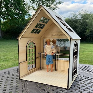 May include: A wooden dollhouse dance studio with a figure inside. The studio has arched windows, a sloped roof, and the text "One's Dance Studio". The doll is wearing a cream-colored shirt and blue shorts. The studio is on a gray table.