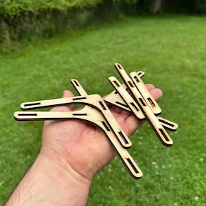 May include: A collection of unfinished wooden shelf brackets with a natural wood grain. The brackets have a unique, angular design with cut-out details. The items are held in a hand against a backdrop of green grass.