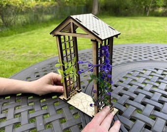 Puede incluir: Un enrejado de jardín de madera en miniatura con un diseño de celosía, un techo de tejas y una base con un patrón de piedra. El enrejado está adornado con flores moradas y hojas verdes. La escena está ambientada al aire libre sobre una mesa.