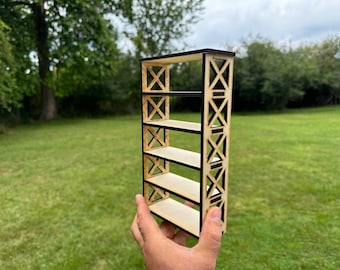 Puede incluir: Una pequeña estantería de madera con cinco estantes. El marco es de color madera clara con ribetes negros y cortes decorativos en forma de X en los laterales. Los estantes son de color madera clara. La estantería se sostiene frente a un fondo verde y herboso.