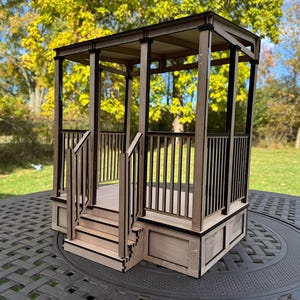 May include: A miniature wooden gazebo with a light brown frame and black accents. The gazebo features a slatted railing, a small set of stairs, and a flat roof. It is set on a dark gray table outdoors.
