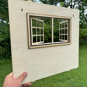 May include: A wooden panel with a laser-cut window design. The window frame is a darker shade of brown, with two window panes that open outwards. The panel is held against a background of green foliage and grass.