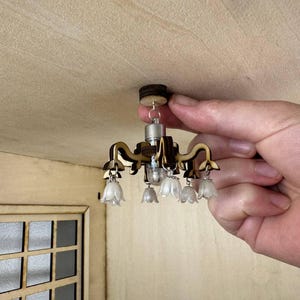 Puede incluir: Candelabro en miniatura con marco marrón oscuro y seis luces blancas en forma de tulipán. El candelabro está sostenido por una mano, mostrando su pequeño tamaño. El accesorio está diseñado para una casa de muñecas o un entorno en miniatura similar.