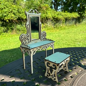 May include: A miniature wooden vanity set with a mirror, table, and stool. The set features intricate cut-out designs and a light blue tabletop. The mirror is rectangular, and the stool is small and square. The set is displayed outdoors.