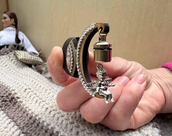 Op de afbeelding: Een decoratieve zwart-zilveren armband met een zilveren engelbedel en een kleine zilveren lamp. De armband is versierd met heldere strass-steentjes. De achtergrond omvat een pop en een gehaakte deken.