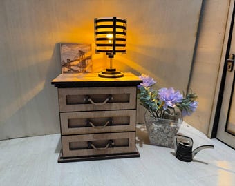 May include: A miniature wooden nightstand with three drawers, a small lamp, and a framed picture. The lamp has a cylindrical shade with horizontal slats, casting a warm glow. A vase of purple flowers and a small watering can are also present.