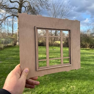 May include: A wooden window frame with a rectangular design. The frame is a light brown color and features a grid pattern within the window space. The background shows a green grassy area and trees.