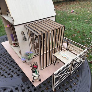 Op de afbeelding: Een miniatuur houten poppenhuis met een lichte houten afwerking. Het huis heeft een veranda met een tafel en stoelen, een balkon en een kleine tuin. De details omvatten een raam, deur en decoratieve elementen. Het poppenhuis staat op een donker oppervlak.