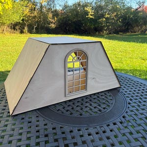 May include: A wooden dollhouse with an arched window. The structure is light brown with a dark trim, and sits on a dark metal table. The background features green grass and trees under a sunny sky.