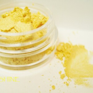 May include: A small, clear jar filled with bright yellow powder, with some spilled beside it. The word "SUNSHINE" is written in yellow at the bottom. The product appears to be a cosmetic, possibly eyeshadow or highlighter.