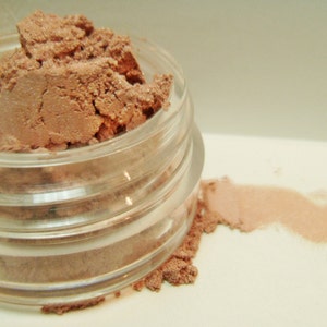 May include: A clear glass jar filled with a light brown, powdery substance, possibly eyeshadow or pigment. A small amount of the powder is spilled out, creating a streak of color. The word "Flesch" is written in orange script.