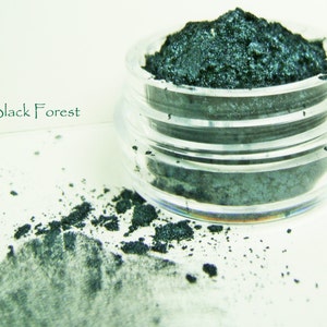 May include: A clear, small jar filled with dark green powder, with some spilled on the surface. The text "Black Forest" is written in green cursive on the left side of the image. The powder has a slightly shimmery appearance.