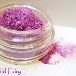 May include: A small glass jar filled with a vibrant purple shimmery eyeshadow. The eyeshadow is spilling out of the jar onto a white surface.