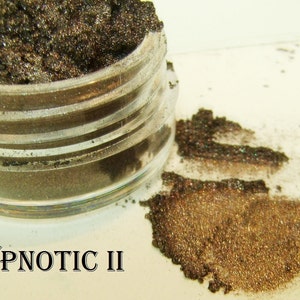 Könnte beinhalten: Ein kleines, klares Glas mit dunklem, schimmerndem Lidschatten. Ein kleiner Haufen des gleichen Lidschattens befindet sich neben dem Glas. Der Text "HYPNOTIC II" ist darunter gedruckt.