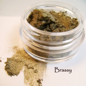 Pode incluir: Um pequeno frasco de vidro cheio de pigmento de sombra de olhos marrom dourado. O frasco está aberto e parte do pigmento caiu na superfície branca. O texto "Brassy" está escrito abaixo do frasco.