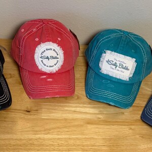 May include: Four distressed baseball caps in black, red, teal, and navy blue. Each cap features a frayed-edge patch with the text "The Salt Runs Deep In Her Soul" and "Salty Bitch" in blue script, along with white contrast stitching and visor wear.