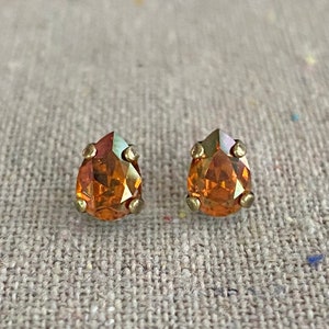 May include: A pair of teardrop-shaped stud earrings. The earrings feature orange-colored crystals with a gold-colored metal setting. The crystals have a faceted cut, and the earrings are displayed on a textured, neutral-colored surface.