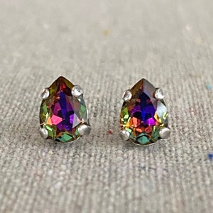 May include: A pair of teardrop-shaped stud earrings. The earrings feature iridescent, multi-colored stones with a silver-colored metal setting. The stones display hues of purple, green, and gold. The earrings are displayed on a textured, light gray surface.