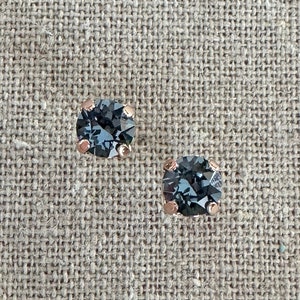 May include: A pair of stud earrings with a round, faceted, dark blue gemstone set in a rose gold-colored metal. The earrings are displayed on a textured, neutral-colored surface.