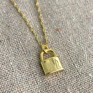 May include: A gold-colored necklace with a delicate chain and a small padlock pendant. The pendant features the engraved initials "JM". The necklace is displayed on a neutral-toned, textured surface.