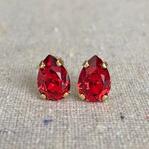 May include: A pair of red teardrop-shaped stud earrings with gold-colored settings. The earrings are set against a neutral, textured background. The red stones have a faceted appearance, catching the light.