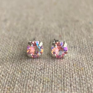 May include: A pair of square-cut stud earrings with iridescent pink and peach-colored crystals. The earrings are set in a silver-colored metal and are displayed on a neutral-toned fabric background. These earrings are a jewelry accessory.