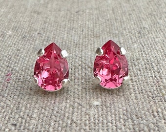 May include: A pair of pink teardrop-shaped stud earrings. Each earring features a faceted, vibrant pink gemstone set in a silver-colored metal bezel. The earrings are displayed on a neutral, textured fabric background.