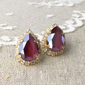May include: A pair of teardrop-shaped stud earrings. Each earring features a large, faceted, burgundy-colored stone surrounded by a halo of small, clear crystals. The setting is gold-toned, and the earrings are displayed on a textured, neutral-colored surface.
