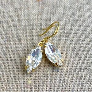 May include: A pair of gold-toned earrings featuring clear, marquise-cut crystals. The crystals are set in a gold bezel and hang from gold-colored ear wires. The earrings are displayed on a textured, neutral-colored surface.