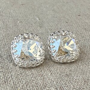 May include: A pair of square-shaped stud earrings with a clear, faceted crystal center. The crystals are surrounded by a silver-colored, ornate filigree setting. The earrings are displayed on a neutral-colored fabric background.