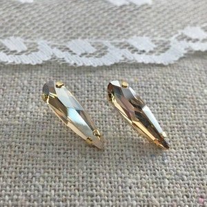 Puede incluir: Dos cristales facetados en forma de lágrima, de color marrón claro, con engastes de metal dorado. Se muestran sobre una superficie texturizada de color neutro, con un ribete de encaje blanco en el fondo.