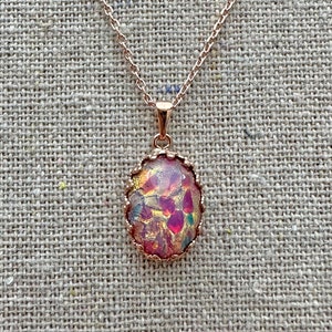 May include: A rose gold necklace with an oval pendant. The pendant features a colorful, iridescent stone with shades of pink, purple, and gold. The stone is set in a decorative, textured frame. The necklace chain is delicate and also rose gold.
