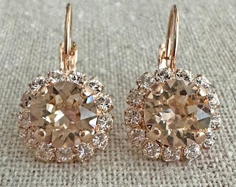 May include: A pair of rose gold leverback earrings featuring a large, round, champagne-colored faceted center stone surrounded by a halo of smaller, clear, round-cut crystals. The metal setting has a warm, metallic rose gold finish.