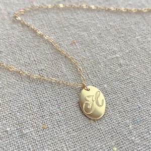 May include: A gold-colored necklace with a delicate chain and an oval pendant. The pendant features a cursive letter "H" engraved in the center. The necklace is displayed on a neutral-toned fabric background.