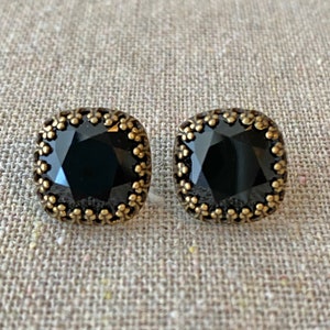 May include: A pair of square black gemstone stud earrings with a gold-colored, ornate border. The earrings have a faceted, dark gemstone center and a decorative, floral-like edge. The earrings are displayed on a neutral, textured surface.