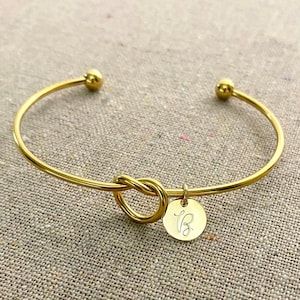 May include: A gold-colored bangle bracelet with a knot design in the center and a small, round charm. The charm is engraved with the letter "B". The bracelet has a simple, elegant design.