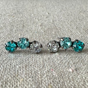 May include: A pair of silver-toned stud earrings. Each earring features three round, sparkling stones: two shades of teal and one clear. The stones are set in a row, creating a subtle, elegant design.