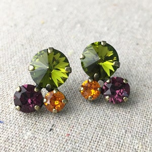 May include: A pair of stud earrings featuring three colorful crystals each. The earrings have a large, round, olive-green crystal at the top, with a smaller orange crystal and a deep purple crystal below. The crystals are set in a gold-tone metal.