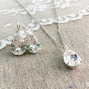 May include: A jewelry set consisting of a silver-toned necklace and a pair of matching leverback earrings. Each piece features a clear, faceted, pear-shaped crystal pendant set in a simple metal prong setting, suspended from a delicate silver chain.
