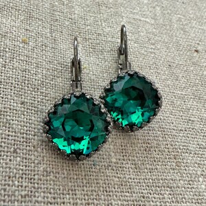 May include: A pair of emerald green square-cut earrings with a dark metal frame and lever-back closures. The earrings have a faceted design, reflecting light. The jewelry is displayed on a neutral-toned surface.