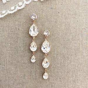 May include: Elegant, rose gold-toned chandelier earrings. Each earring features a large, clear, teardrop-shaped crystal suspended from a smaller, round crystal. Additional smaller teardrop crystals dangle below. The earrings are displayed on a neutral linen background.